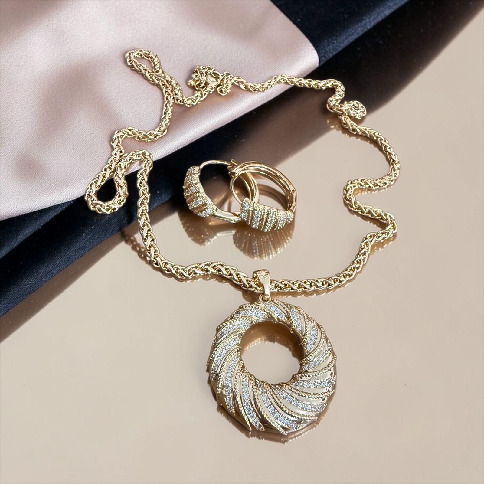 Conjunto Aura Espiral Lux Descrição: Conjunto sofisticado com colar de corrente trabalhada e pingente em formato circular com design espiral cravejado em zircônias, acompanhado de brincos elegantes no mesmo estilo. Uma composição moderna e luminosa, perfeita para quem busca presença, movimento e brilho em cada detalhe.